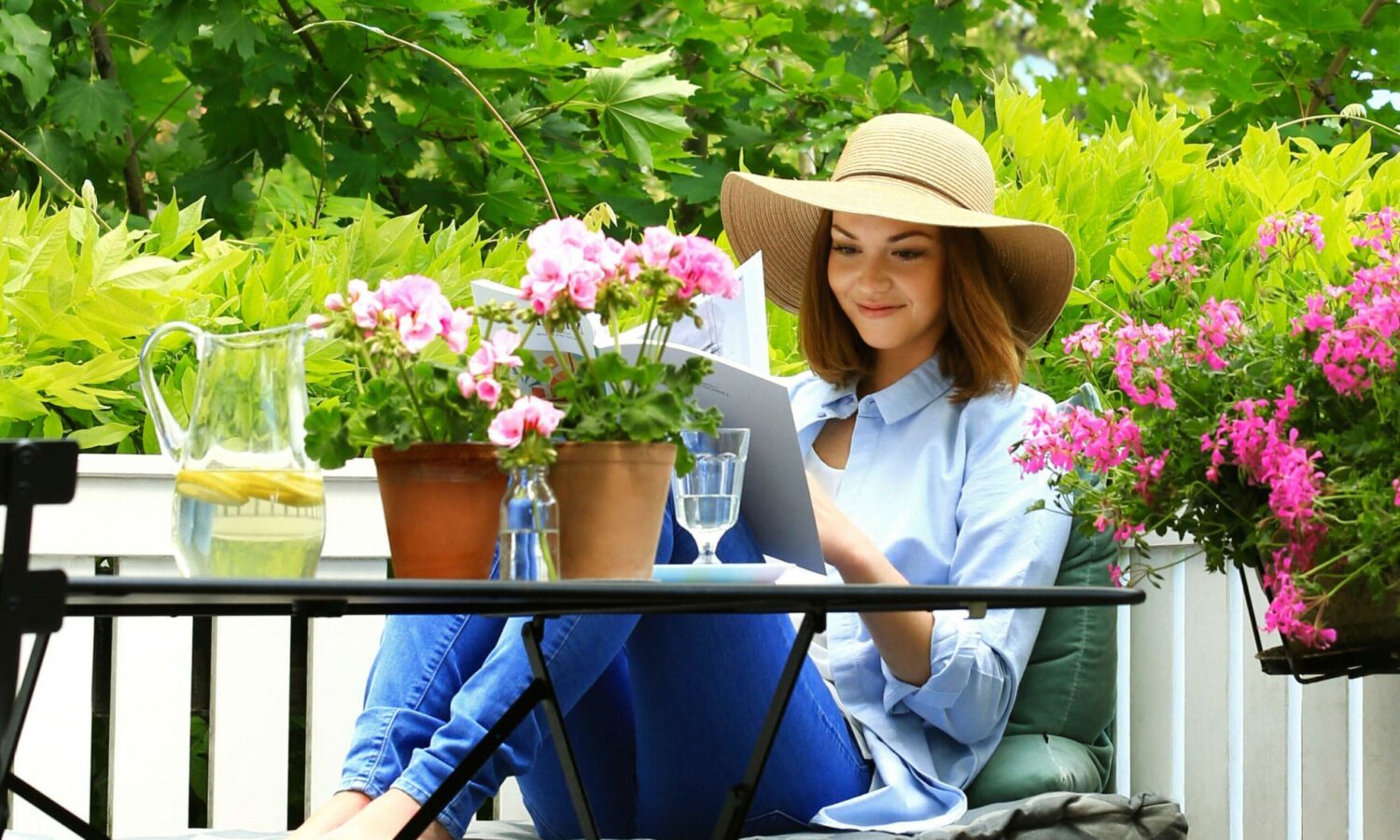 Frau mit Flechthut sietzt auf Bank mit Zeitung in der Hand. Neben ihr ein Tisch mit rosa Geranien in Tontöpfen. Hinter ihr pinke Geranien in Pflanzkasten an hölzernem Balkongeländer.